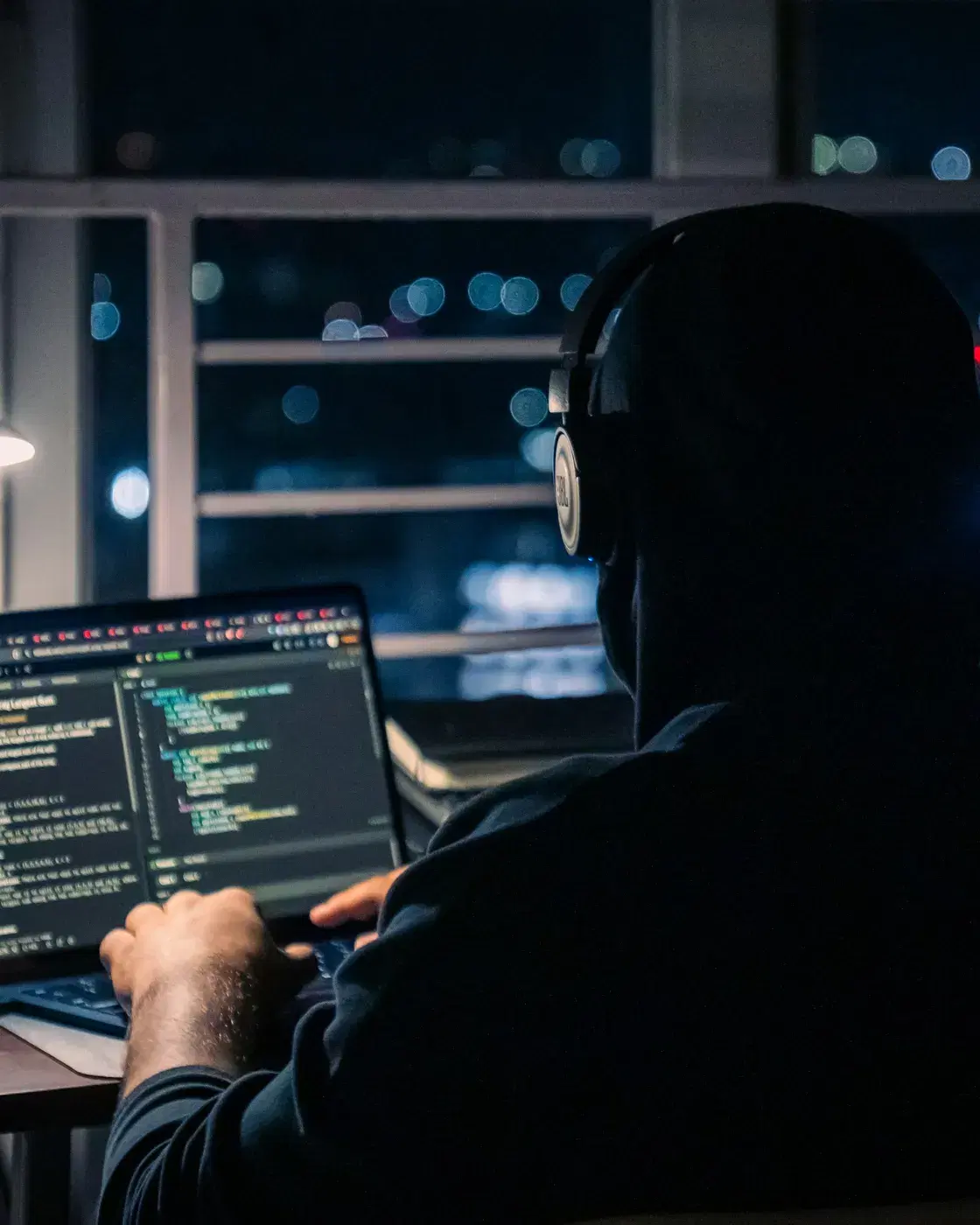 Someone in headphones, seen from behind, writing code on a laptop at night
